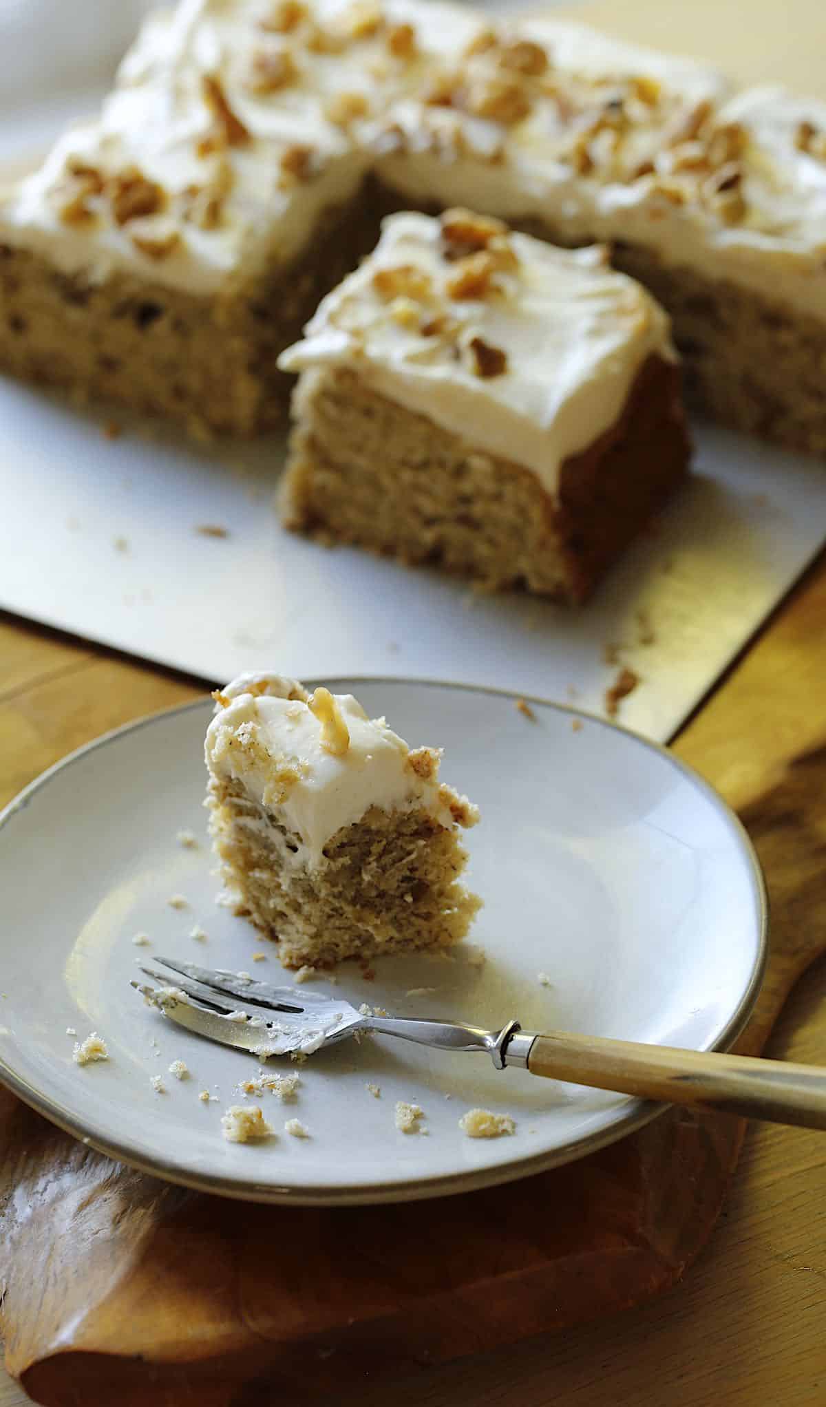 A piece of banana cake with cream cheese frosting on a small plate with fork, whole cake sliced in background