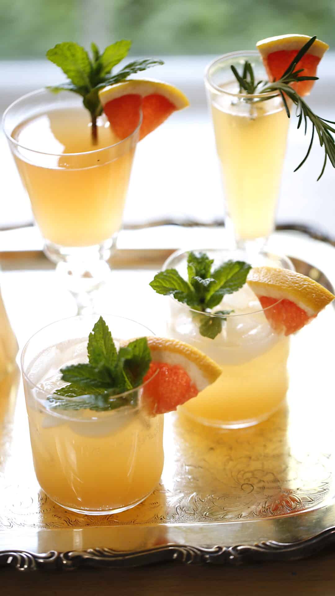 a vertical image of four cocktails on a tray garnished with fruit and herbs