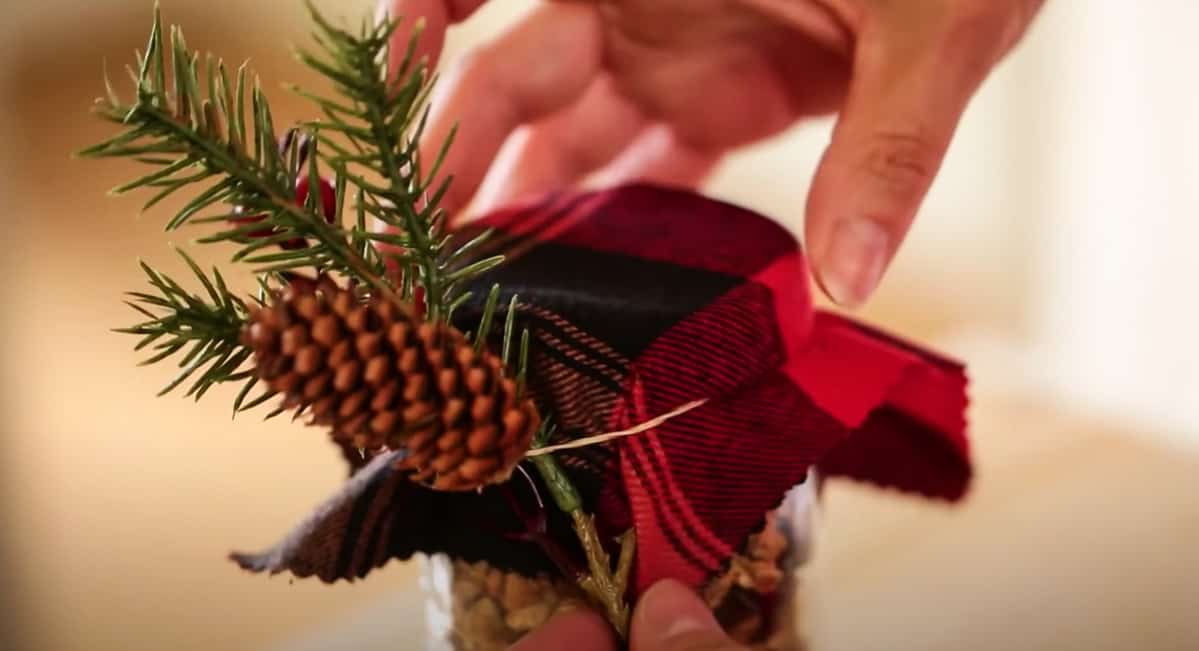 adding faux evergreen to the top of a mason jar covered with red check fabric
