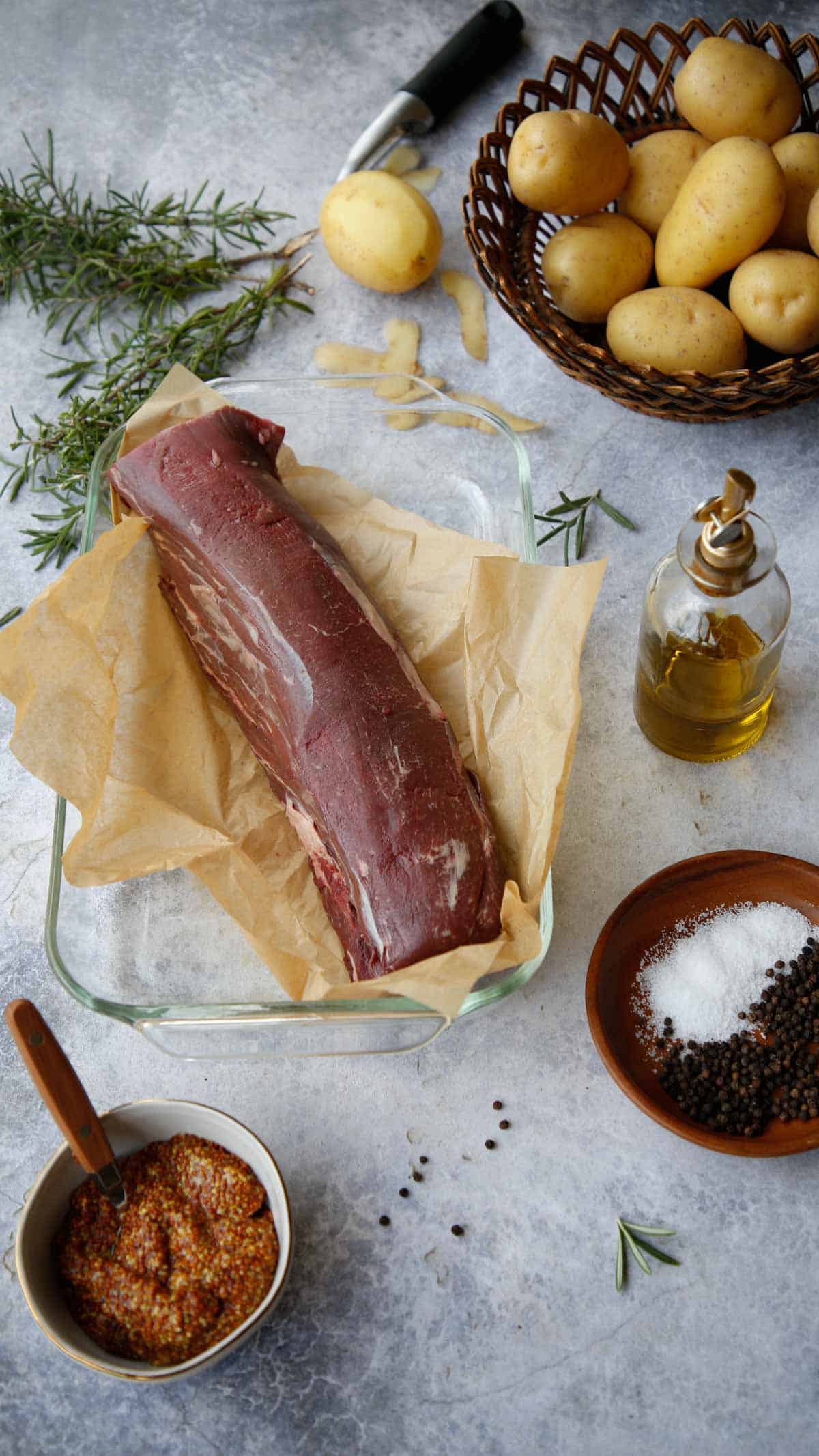 Ingredients on a counter including a tenderloin of beef, salt, potatoes, olive oil, rosemary and mustard