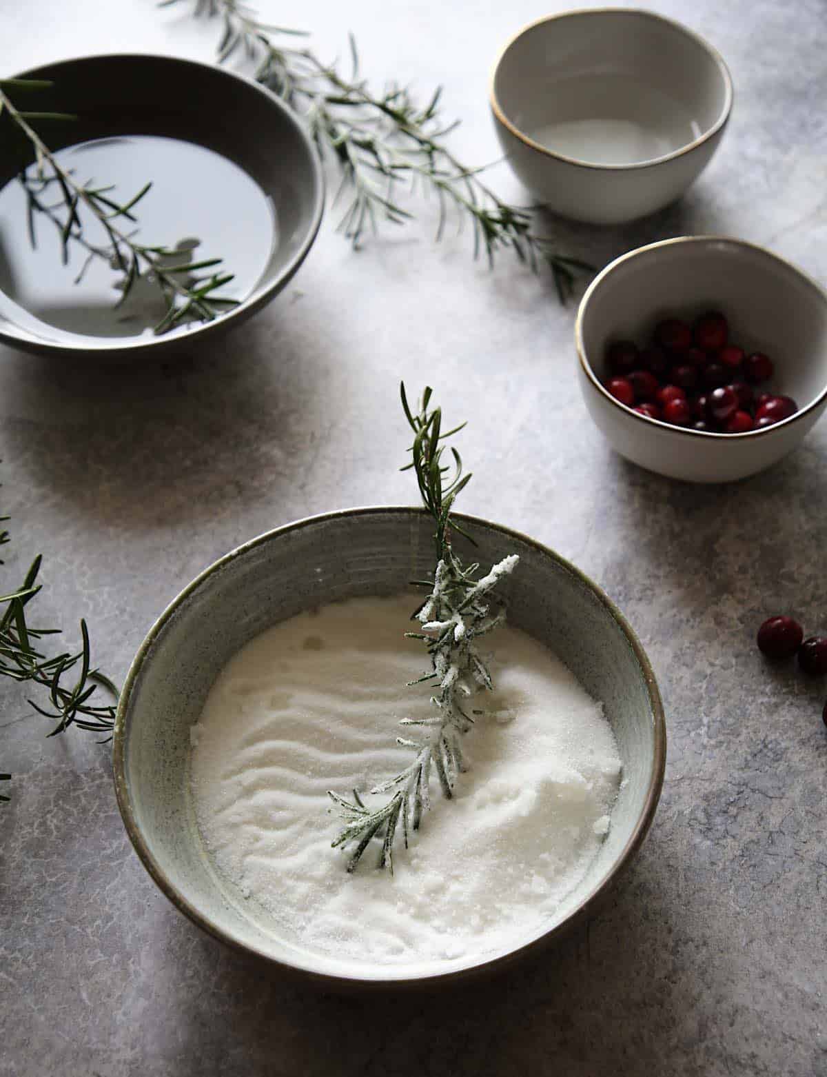 rosemary sprigs dipped in bowls of water and sugar along with cranberries