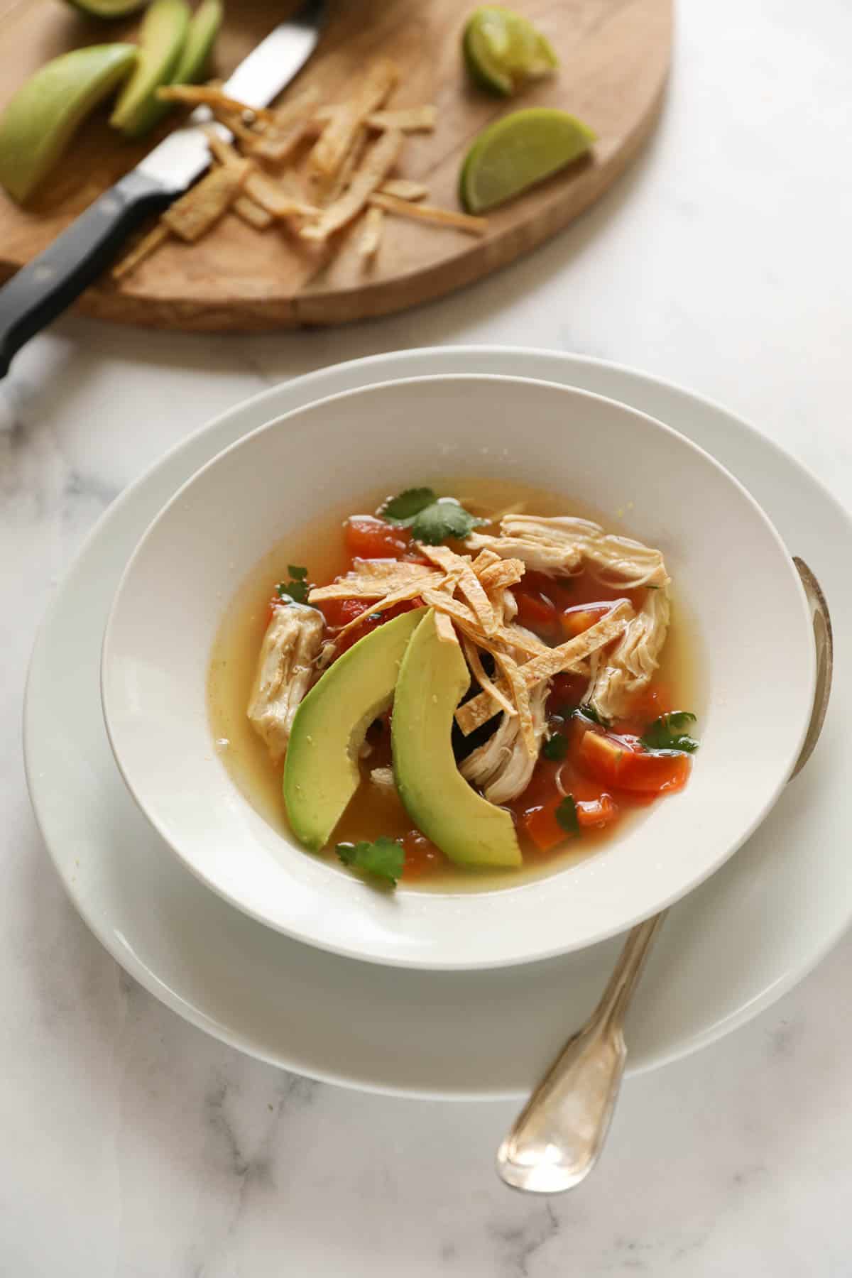 A bowl of soup with chicken, tortillas and avocado