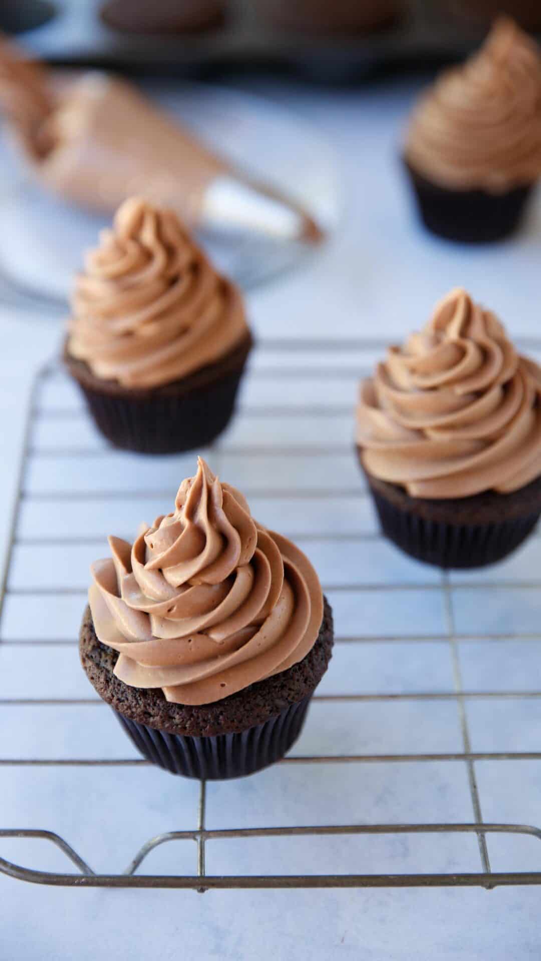 Chocolate cupcakes being frosted with chocolate frosting on a cooling rack
