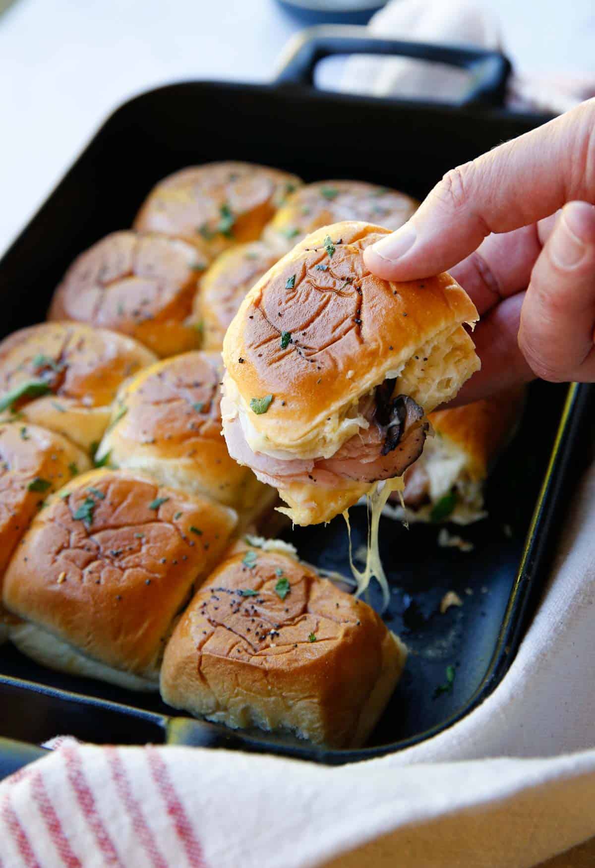 a man's hand pulling apart a slider to show the filling of ham, and gooey cheese
