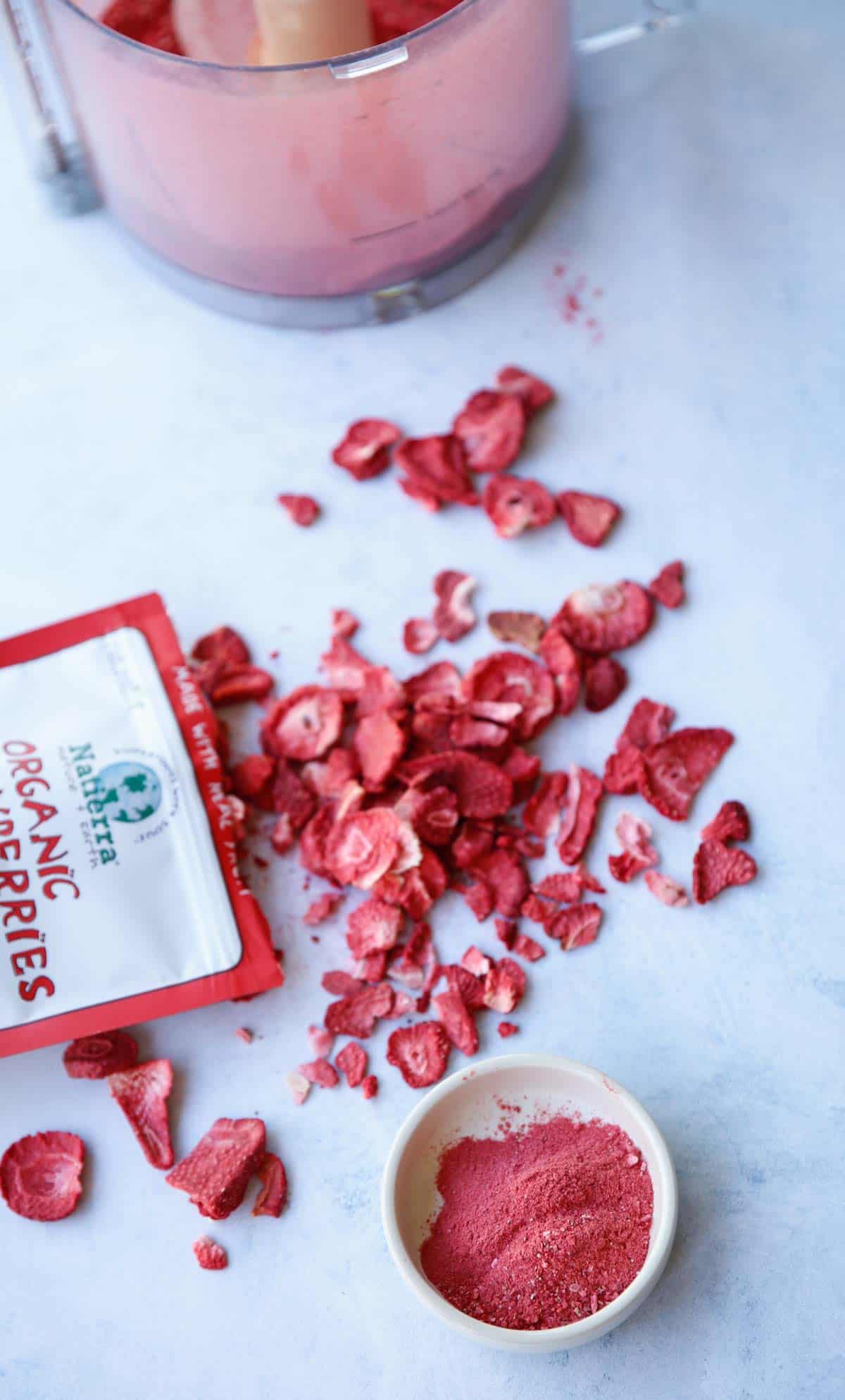 Dehydrated Strawberries coming out of a package with a food processor bowl in the background and ground strawberry dust in the foreground