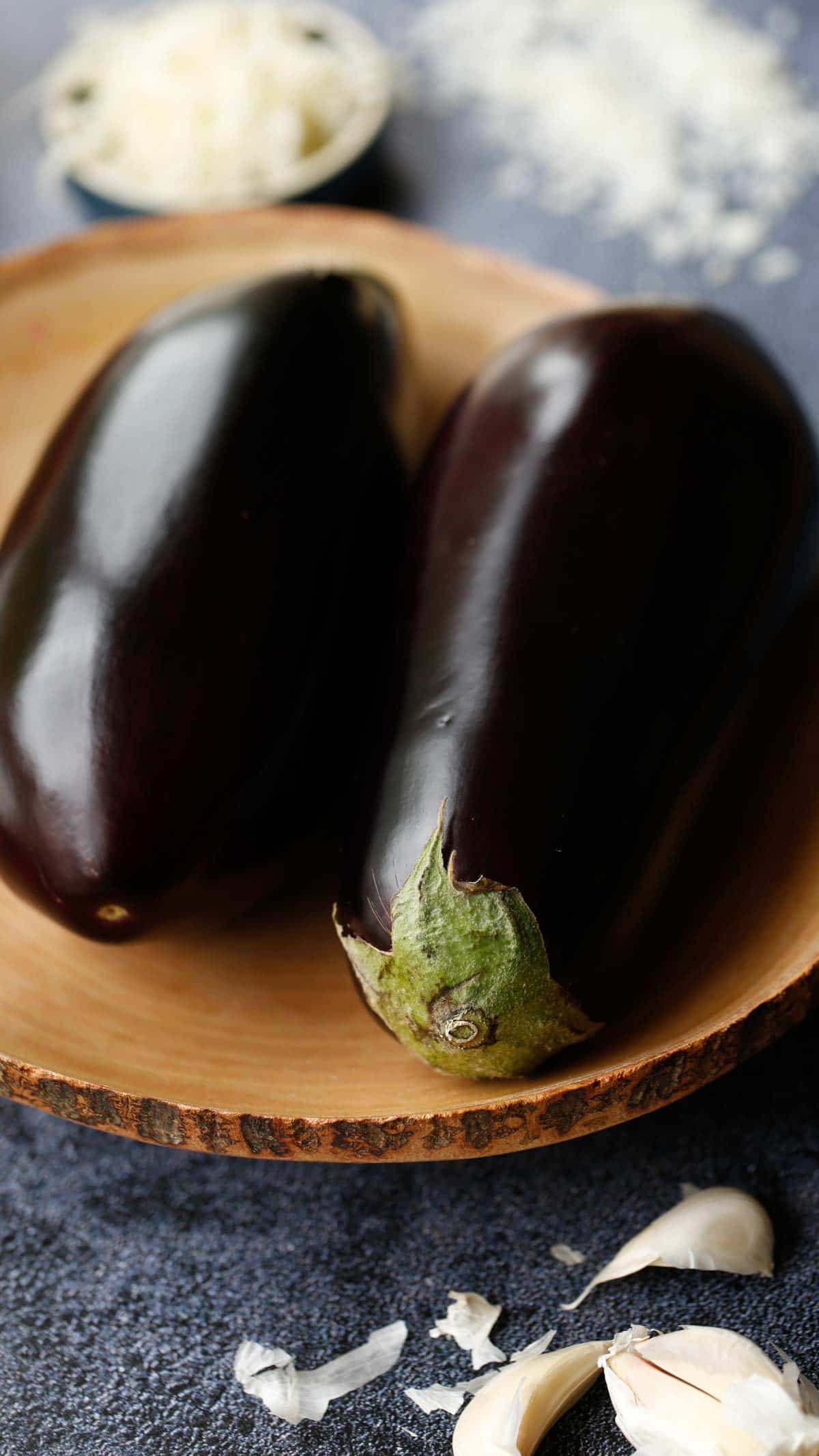 2 eggplant in a wooden bowl with garlic and cheese in the background