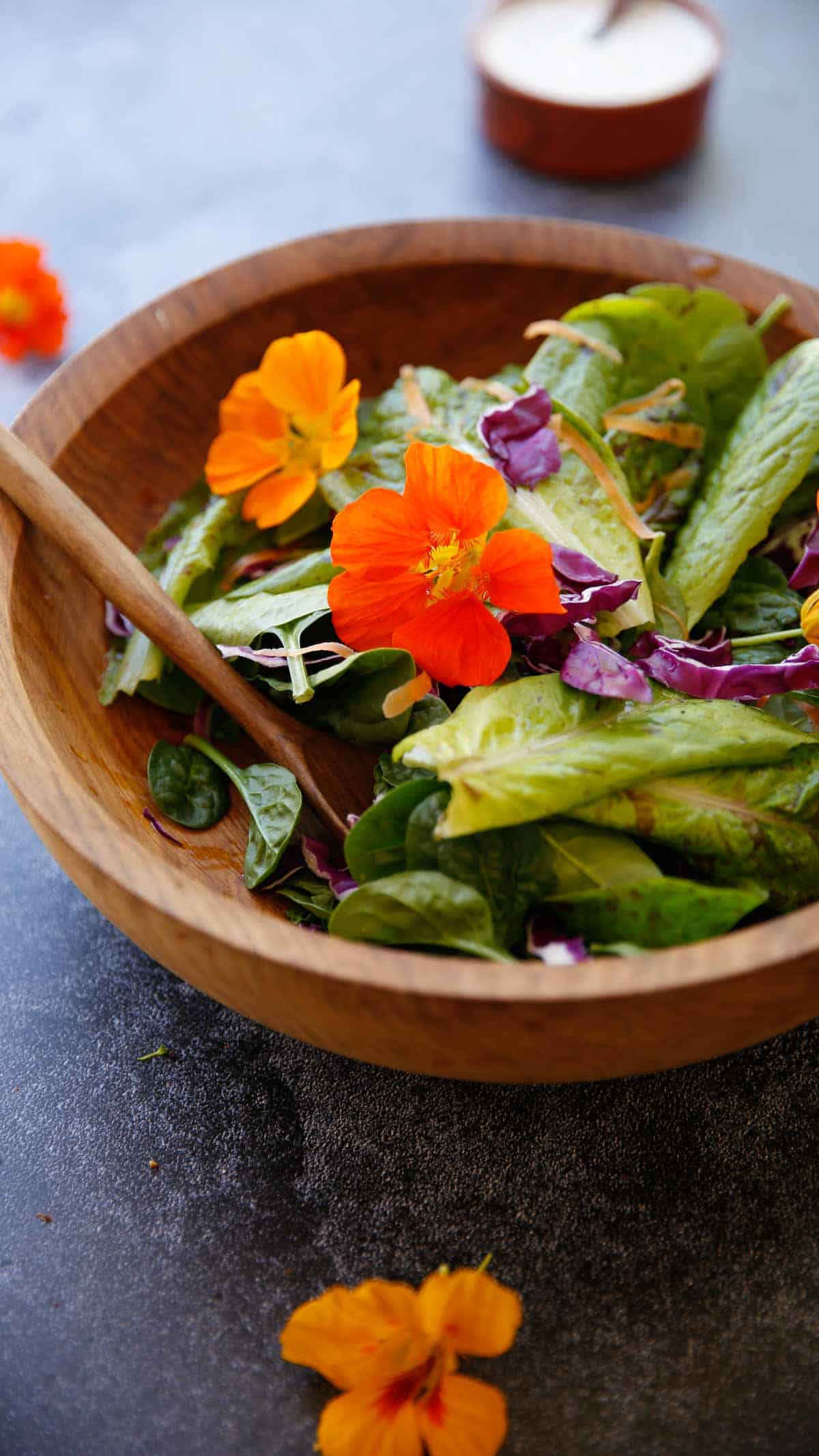 Greens in a salad bowl with edible flowers and a small bowl of dressing in the background