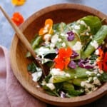 a wooden salad bowl filled with greens and nasturtiums with dressing in the background