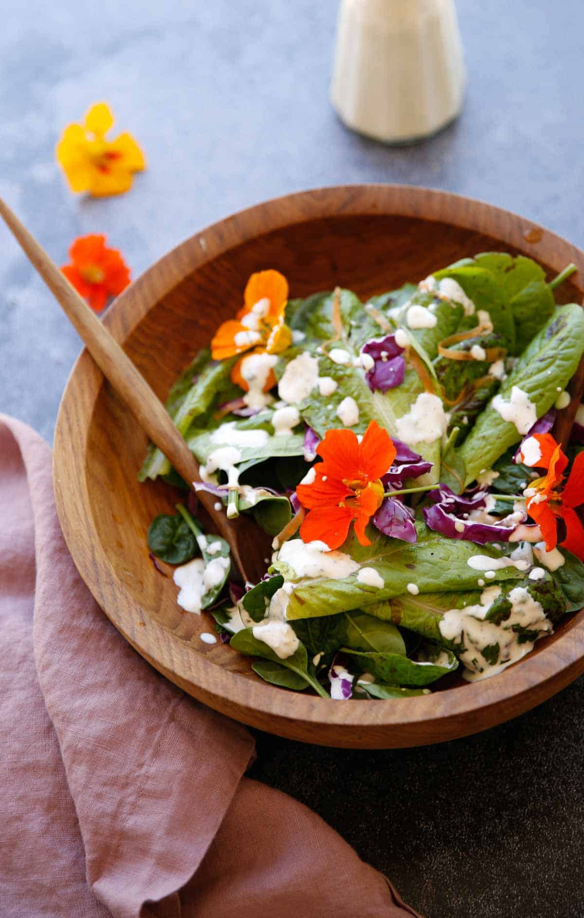 a wooden salad bowl filled with greens and nasturtiums with dressing in the background 