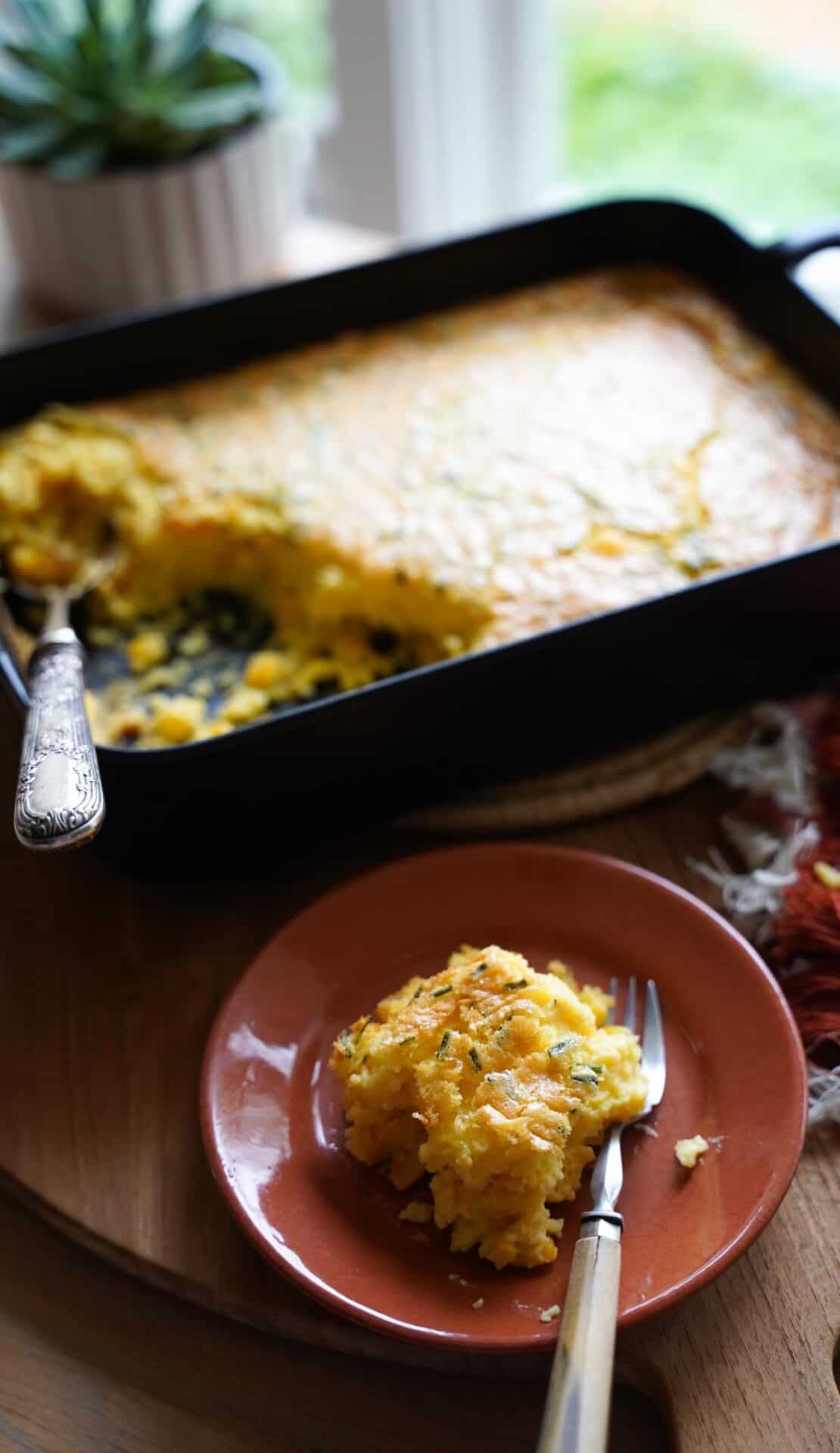 A serving of corn pudding on a plate with a casserole dish in the background