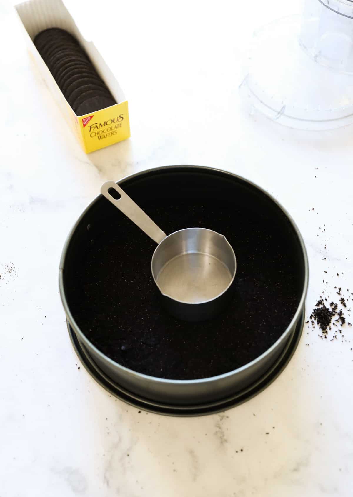 a chocolate cookie crust being fitted into a cheesecake pan with a measuring cup