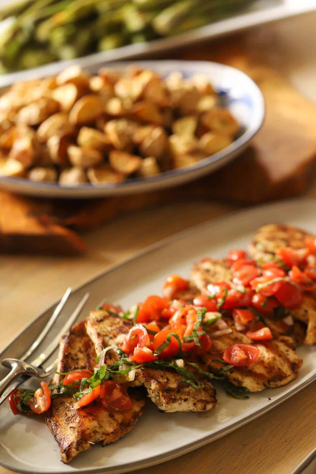 Grilled chicken topping with cherry tomatoes and a platter of potatoes and asparagus in the background