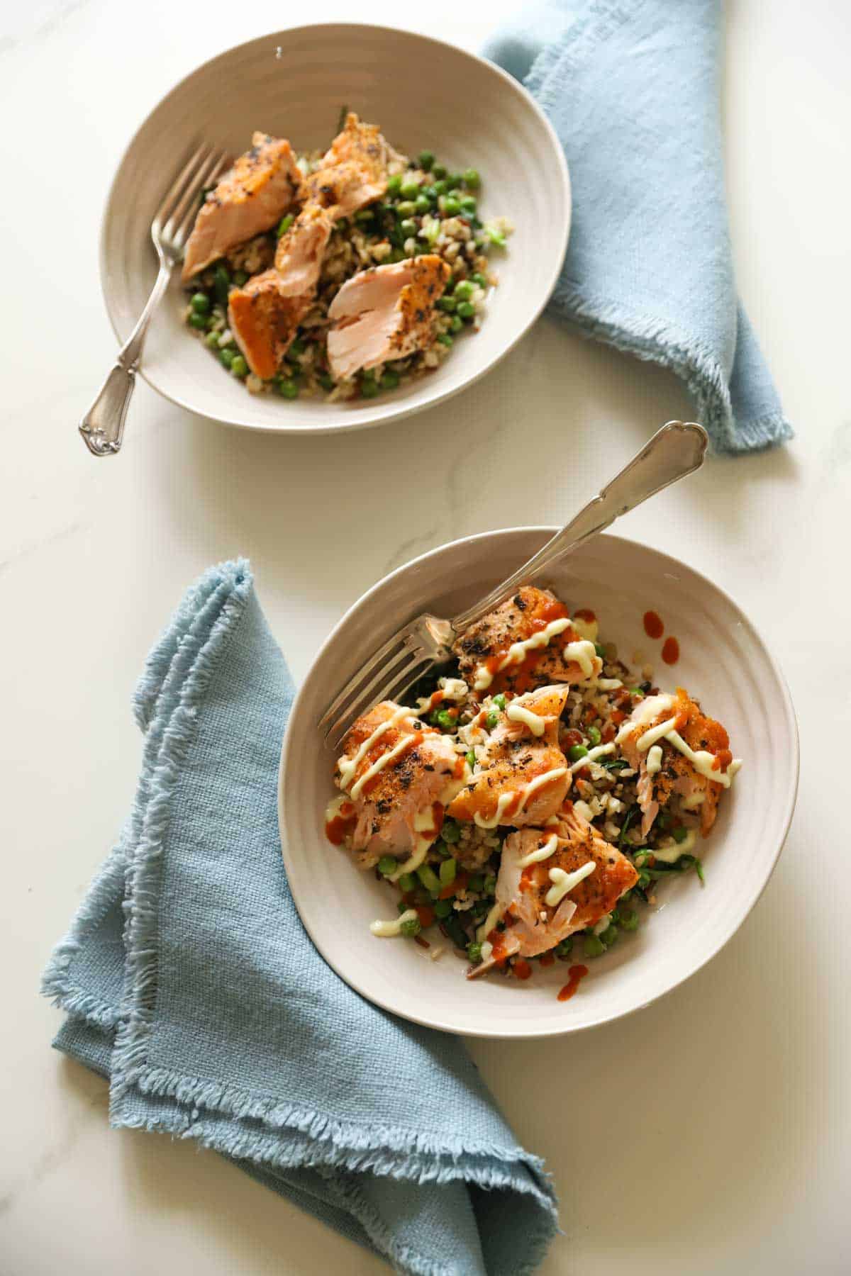 two salmon bowls and rice with blue napkins. One drizzled with Kewpie mayonnaise and sriracha