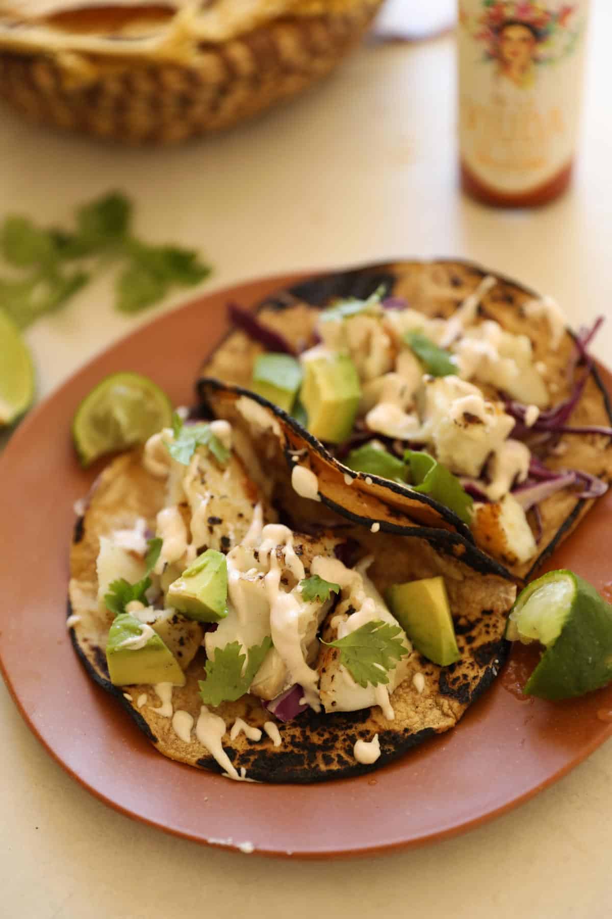 a plate of two fish tacos with avocado, crema Mexicana, cabbage and cilantro