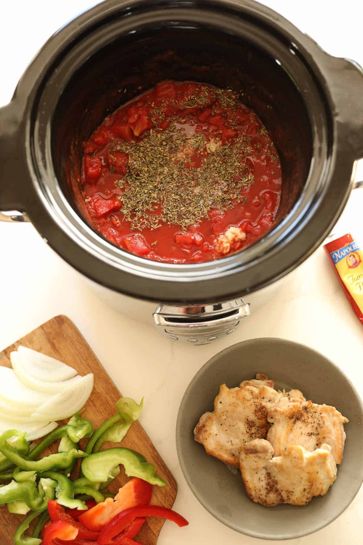 birds eye view looking into a slow cooker with a cacciatore mixture. Seared chicken on a plate with sliced peppers and onions on another plate