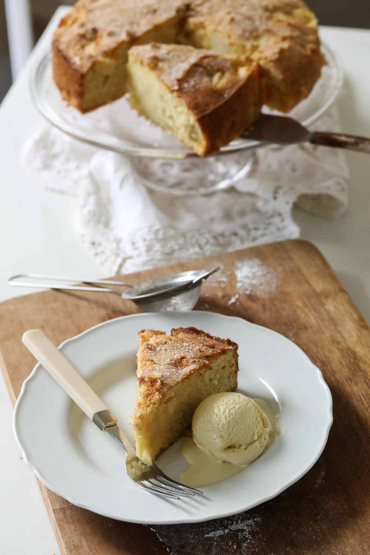 A whole cake on a cake stand with a slice of came on a plate with vanilla ice cream and a fork