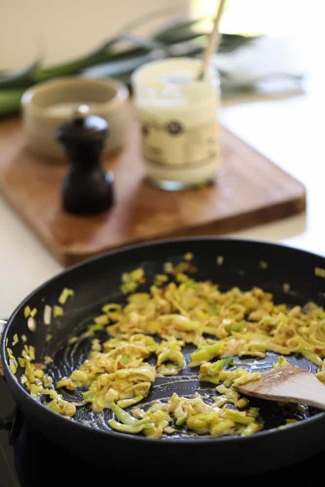 Creamy leeks in a skillet with a cutting board in the background with ingredients on it.