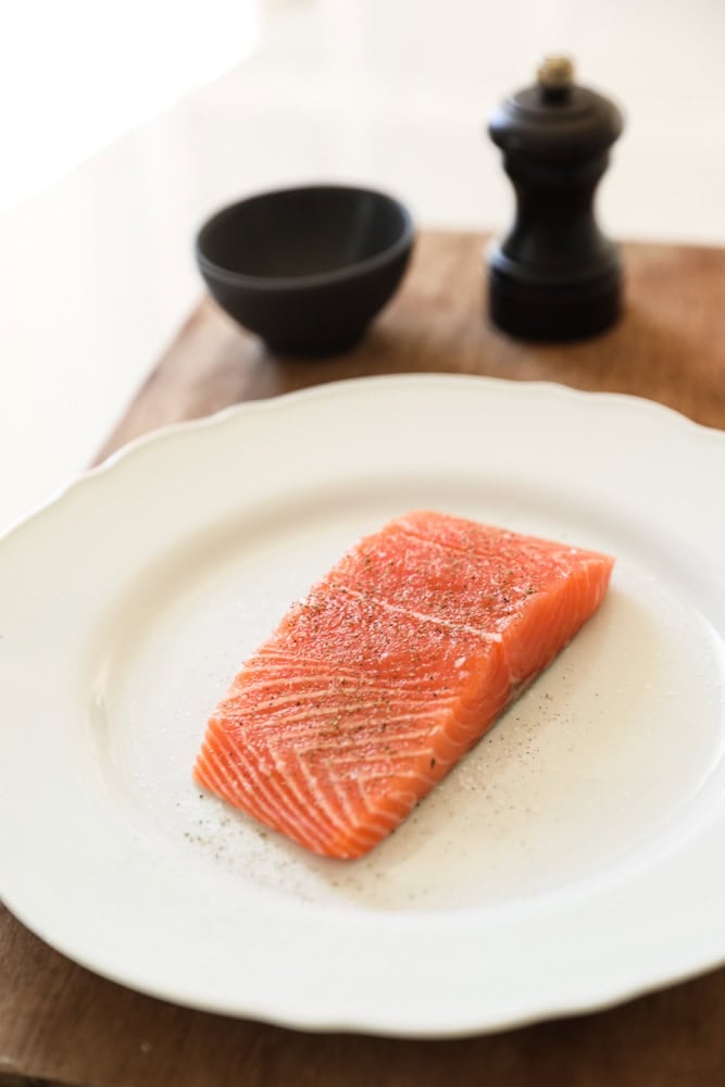 a filet of salmon seasoned with salt and pepper resting on a white plate.