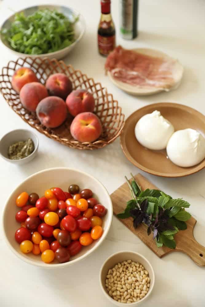 Peaches, Burrata, Rocket, Cherry Tomatoes, Fresh Herbs, Pine Nuts, Prosciutto on a counter top.
