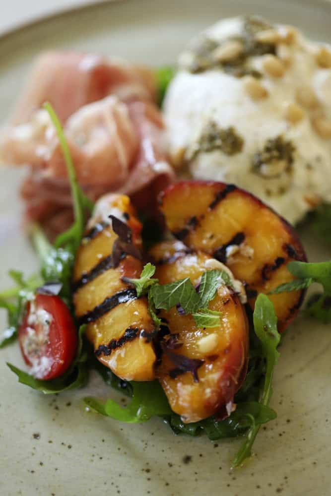 Grilled peaches with an aged balsamic vinaigrette with burrata and prosciutto on a plate.