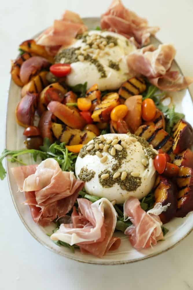 A platter of grilled peaches, arugula, cherry tomatoes and burrata cheese drizzled with pesto sauce.