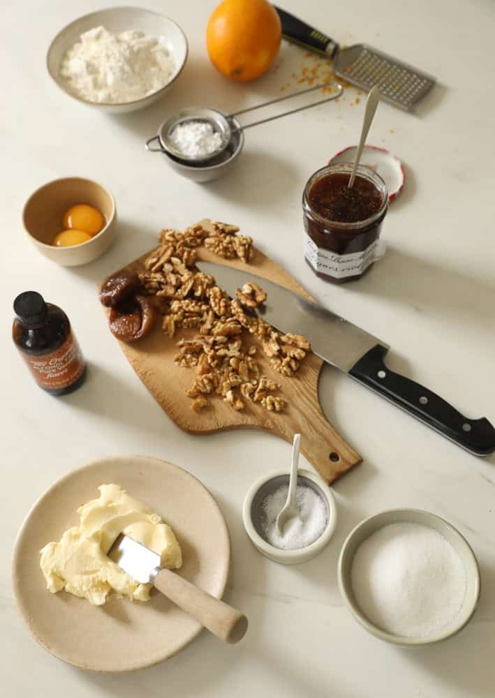 Walnuts, figs, butter, orange, flour and sugar laid out on a countertop.