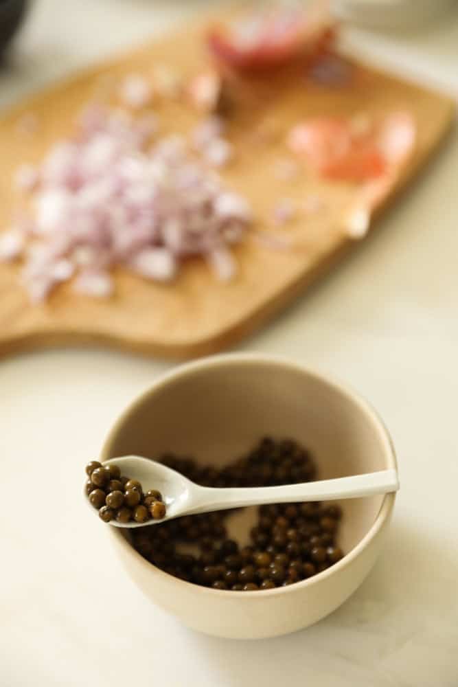 Peppercorns in a small bowl with a small spoon. Diced shallots in the background. 