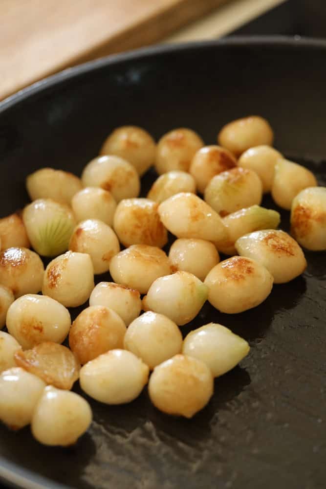 Mini white onions caramelizing in a non-stick pan.