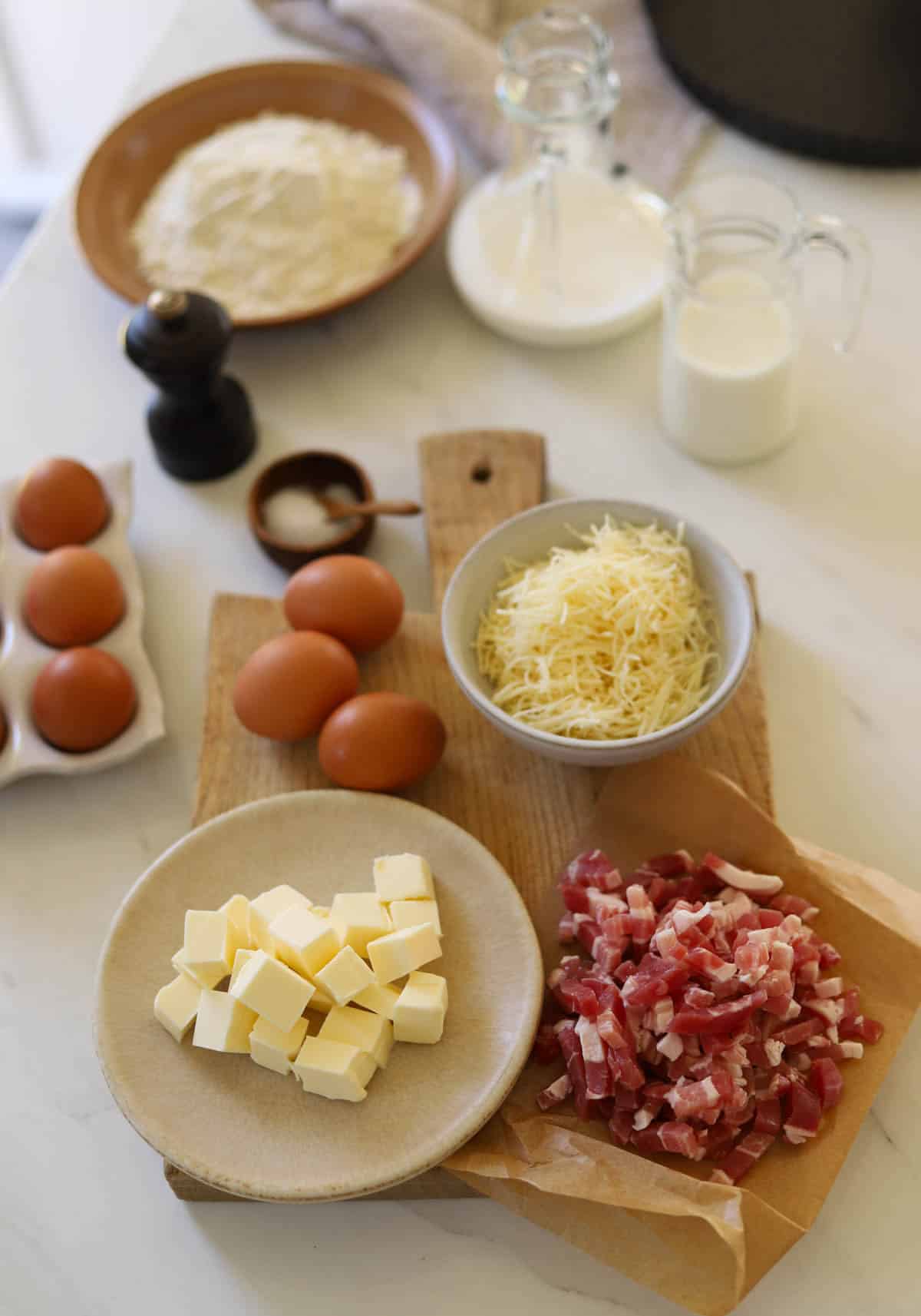 Ingredients laid out on a counter including eggs, cream, milk, flour, bacon and cheese.