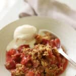 O bowl of strawberry rhubarb crisp with vanilla ice cream and a silver spoon.