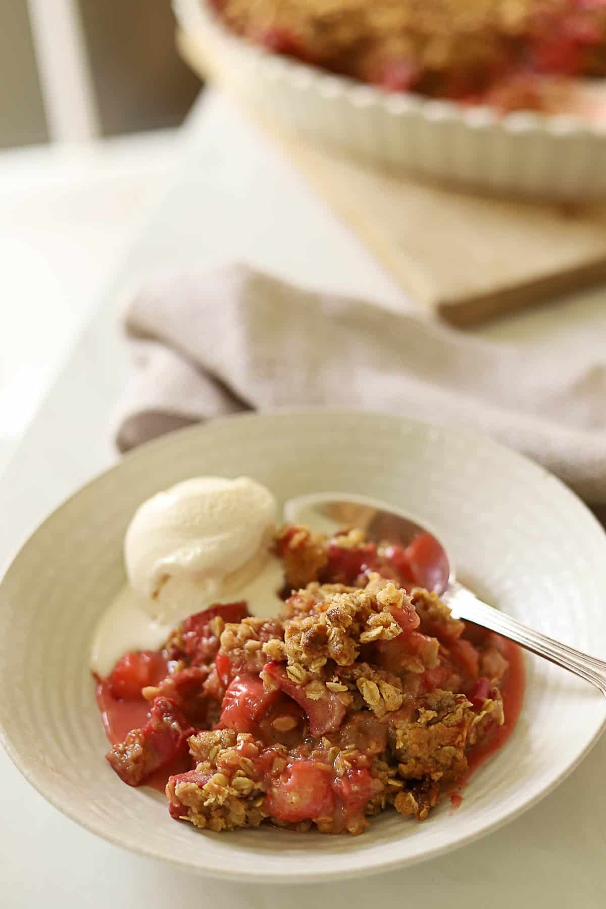 a bowl of fruit crisp with a scoop of vanilla ice cream with the full casserole in the background.